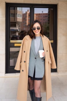 Happy caucasian girl walking with cup of coffee in city on sunny day. Brunette woman wears stylish jacket coat and sunglasses come out from building entrance. Positive emotions lifestyle concept