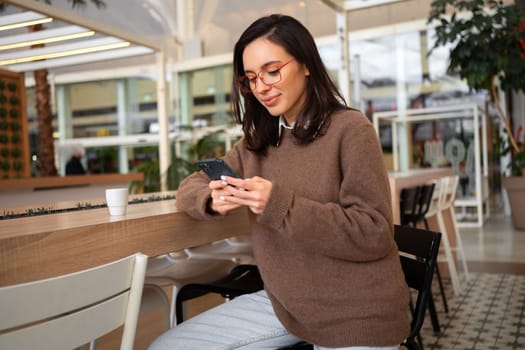 Girl use smart phone look at device screen share social media news wear stylish trendy jumper sitting bar stool. Female smartphone user. Woman in glasses holding smartphone