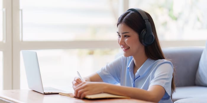 Online education, e-learning. Asian woman in stylish casual clothes, studying using a laptop, listening to online lecture, taking notes, online study at home.