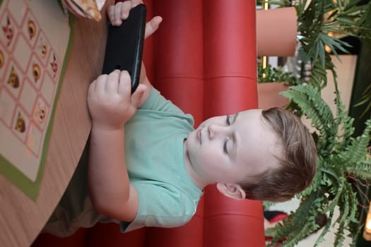 Child, a boy of five years old, is having breakfast at the kitchen table and watching cartoons on his smartphone.