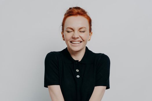 Beautiful and happy pleased red haired woman with closed eyes and toothy smile posing against grey wall. Cheerful excited female demonstrating positive emotions and happiness, dressed casually