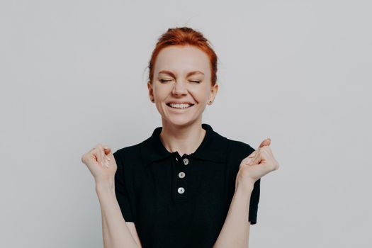 Yes, i did it. Portrait of happy euphoric red haired female with closed eyes holding clenched fists up, excited woman celebrating win, success while posing isolated over grey studio wall