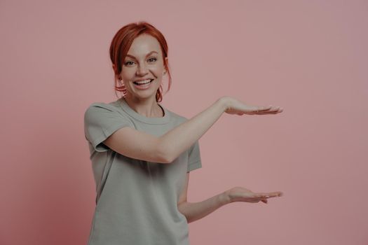 Young pleasant red-haired woman in t-shirt showing huge product size or something big with hands and smiling while standing isolated on pink background. Promotion and advertising concept