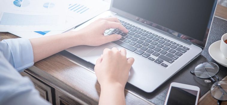 Business concept. Woman in blue shirt typing on computer with coffee on office table, backlighting, sun glare effect, close up, side view, copy space