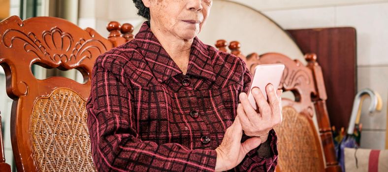 Asian elderly woman sitting and looking through something on modern smartphone, making connection with others at home, living technology, close up