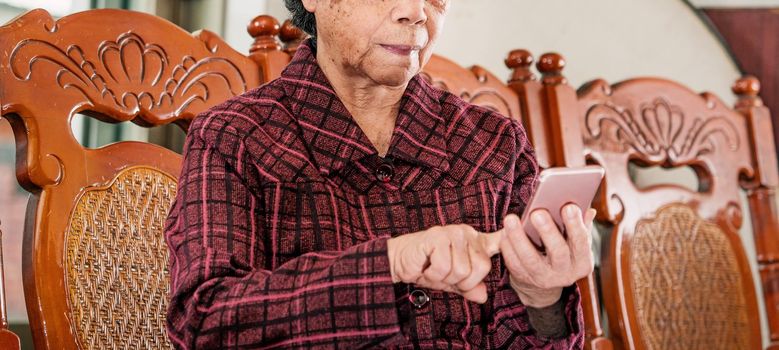 Asian elderly woman sitting and looking through something on modern smartphone, making connection with others at home, living technology, close up