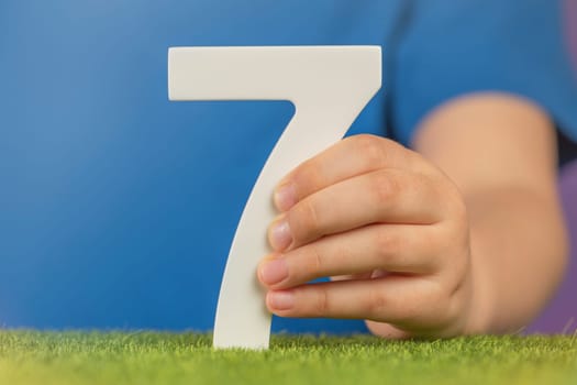 Number 7 in hand. A hand holds a white number seven on green grass and a purple background, seven percent per annum. World Health Day. April 7th. Holiday concept. High quality photo