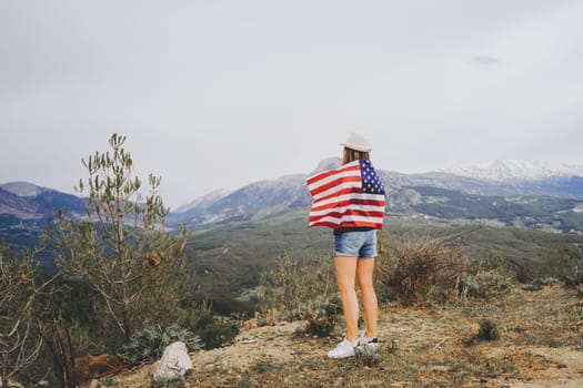 Young woman girl standing on rock cliff with US flag on her back and looking at mountains in background. Female traveller wearing American flag standing mountain top. 4 fourth July Independence Day