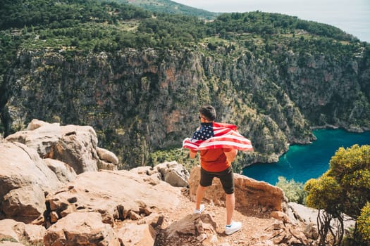 Young man standing on a rock cliff and waving the US flag while looking at sea beneath. Veteran traveller waving the American flag while standing on a mountain top. 4 fourth July Independence Day