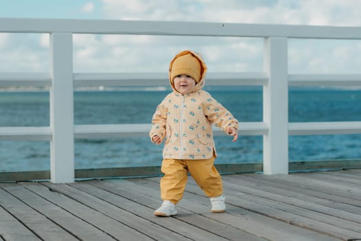 A happy toddler in a yellow jacket and pants strolls on the pier. A baby girl in the hood is having fun near the Baltic Sea.
