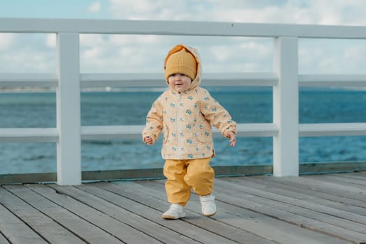 A toddler in a yellow jacket and pants strolls on the pier. A baby girl in the hood is having fun near the Baltic Sea.