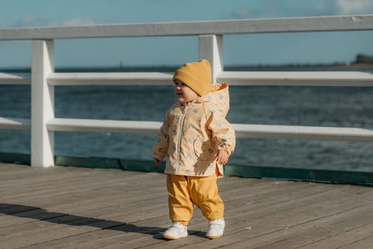 A smiling toddler in a yellow jacket and pants strolls on the pier. A baby girl is having fun near the Baltic Sea.