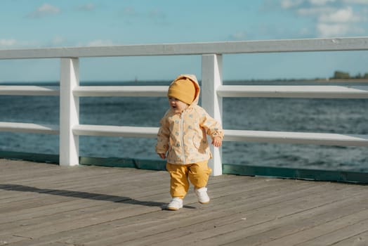 A smiling toddler in a yellow jacket and pants walks on the pier. A baby girl is running and screaming near the Baltic Sea.