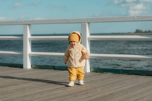 A smiling toddler in a yellow jacket and pants walks on the pier. A baby girl is having fun and screaming near the Baltic Sea.