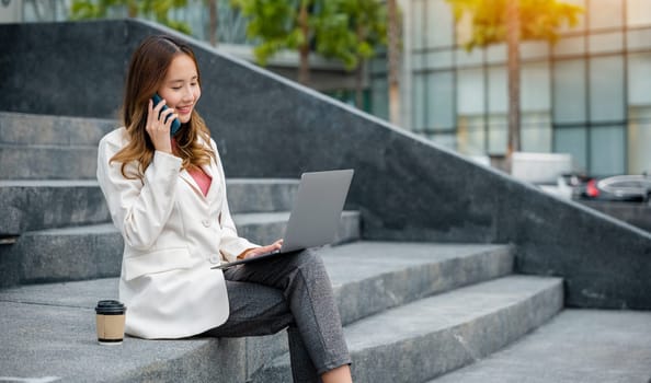 Happy young Asian woman freelance looking laptop with smile while talking on mobile smartphone with coffee cup while sitting on stairs outdoors in the city. Remote working concept in the office.