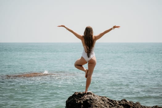 Woman sea yoga. Back view of free calm happy satisfied woman with long hair standing on top rock with yoga position against of sky by the sea. Healthy lifestyle outdoors in nature, fitness concept