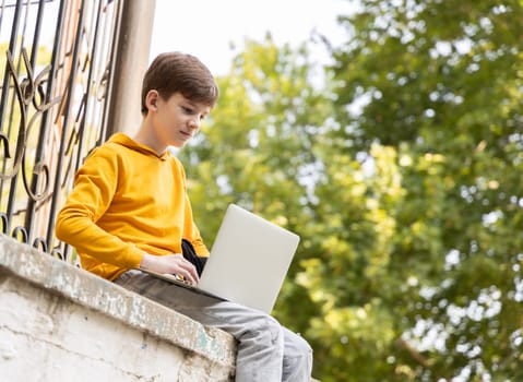 Caucasian child teen boy in yellow hoodie using laptop outdoors. Distant learning on the go. Coding, blogging, surfing, video call