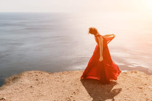 Side view a Young beautiful sensual woman in a red long dress posing on a rock high above the sea during sunrise. Girl on the nature on blue sky background. Fashion photo.