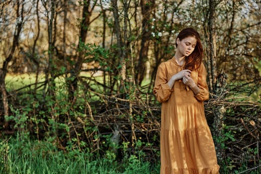 a beautiful woman walks in the shade near the trees, dressed in a long orange dress, enjoying the weather and the weekend, touching her long hair. The theme of privacy with nature, horizontal photography . High quality photo