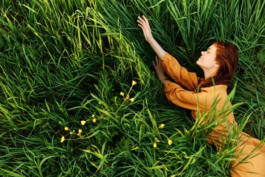 a relaxed red-haired woman enjoys summer lying in the tall green grass in a long orange dress smiling happily looking away. High quality photo