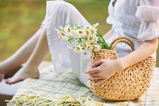 a woman in a light dress sits near the lake and holds a wicker basket with flowers in her hands. High quality photo