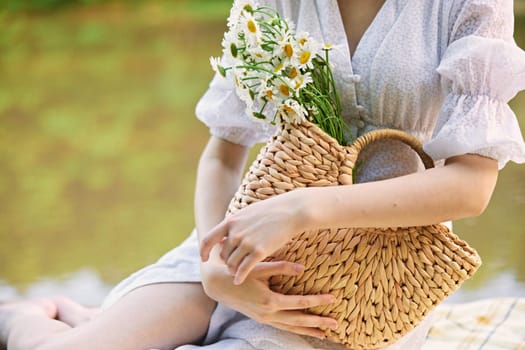 a woman in a light dress sits near the lake and holds a wicker basket with flowers in her hands. High quality photo