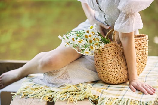 a woman in a light dress sits near the lake and holds a wicker basket with flowers in her hands. High quality photo