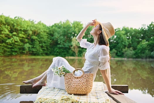 happy woman in a long light dress is resting by the lake sitting on a plaid and enjoying the view. High quality photo