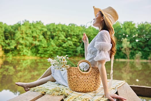 a woman sitting on a pier by the lake in a light dress and a wicker hat smells a chamomile flower. High quality photo