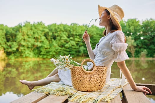 a woman sitting on a pier by the lake in a light dress and a wicker hat smells a chamomile flower. High quality photo