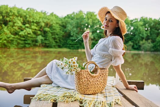 a woman sitting on a pier by the lake in a light dress and a wicker hat smells a chamomile flower. High quality photo