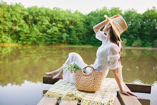 happy woman in a long light dress is resting by the lake sitting on a plaid and enjoying the view. High quality photo