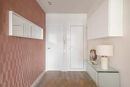 Cozy entrance hall in light colors with an intercom, glass console and a wall cabinet with stylish light and an elongated mirror on peach-colored wall. Concept of a simple laconic room design.