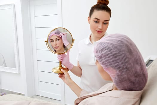 Happy young woman sitting in medical chair and looking in the mirror. She is satisfied after successful beauty treatment with hyaluronic acid fillers