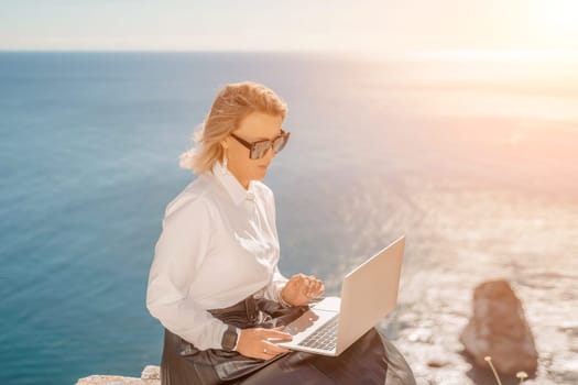 Business woman on nature in white shirt and black skirt. She works with an iPad in the open air with a beautiful view of the sea. The concept of remote work