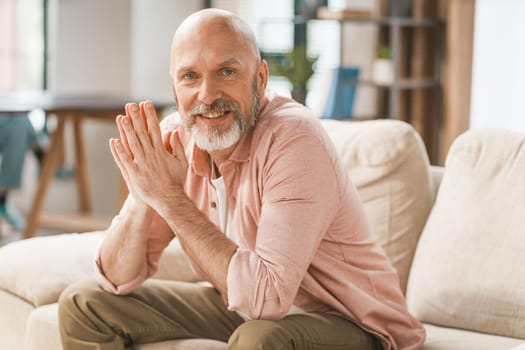 Smiling, happy senior man sitting on a sofa in the comfort of his home. With his hands held together, he radiates a sense of contentment and relaxation. . High quality photo