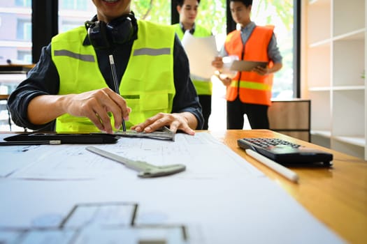 Architects man wearing reflective jacket working with blueprints at office desk with his colleagues standing in background.