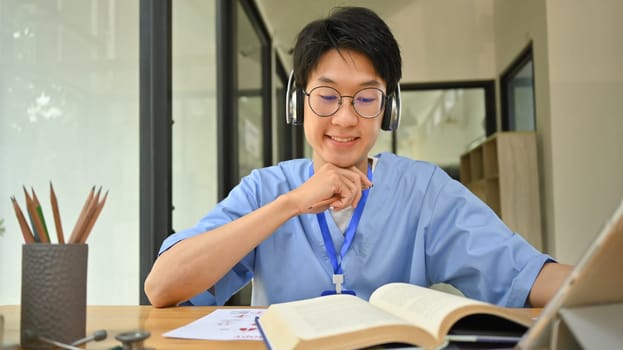 Asian male medical student in headphone reading books, preparing for university exams in library. Medical internship concept.