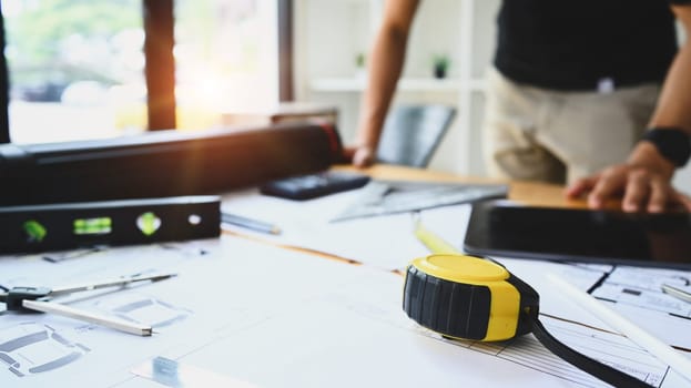 Closeup view of tape measure, blueprints, spirit level and digital tablet on working desk with architect man standing in background.