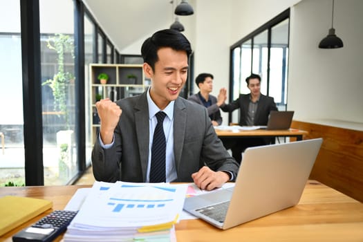 Excited asian male office worker looking at laptop screen, making winner yes gesture, celebrating business success.