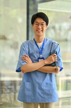 Portrait of young surgeon wearing blue scrubs holding stethoscope standing in hospital. Healthcare and medical concept.