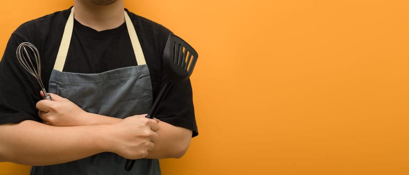 Man in apron holding whisks and kitchen spatula standing against yellow background with copy space.