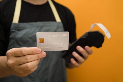 Man wearing apron holding credit card and payment terminal on yellow background. Electronic money, contactless payment concept.