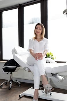 Portrait of Charming Beautician in White Lab Coat Looking at Camera With Teeth Smile, Sitting on Couch, Looking at Camera in Spa Salon. Vertical Plane. High quality photo