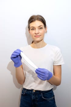 Caucasian manicurist, nail technician holds file in hands, wearing gloves. White smiling manicure master stands near gray wall in spa salon. Soft, safe beauty nail care, treatment. Vertical