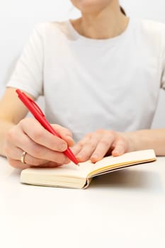 Hands of white woman make note in notebook with red pen. Cropped Caucasian female wears white shirt and marriage ring on hand, writes in a book sitting on chair at white table. Vertical