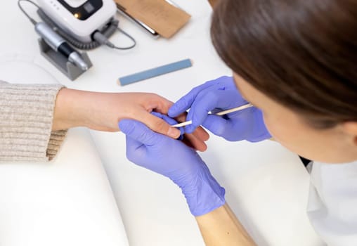 Cropped Nail beautician removes cuticle of female client with orange stick, top view. File, sterilized manicure tools in craft package on white table. Manicure master works in purple gloves.Horizontal