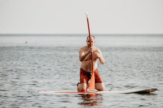 Active mature male paddler with his paddleboard and paddle on a sea at summer. Happy senior man stands with a SUP board. Stand up paddle boarding - outdor active recreation in nature