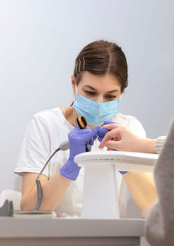 White nail technician in mask drills, removes client's cuticle with hardware machine, holding nail brush. Customer wears warm cloth, sweater. Combined manicure, beauty treatment in spa. Vertical plane