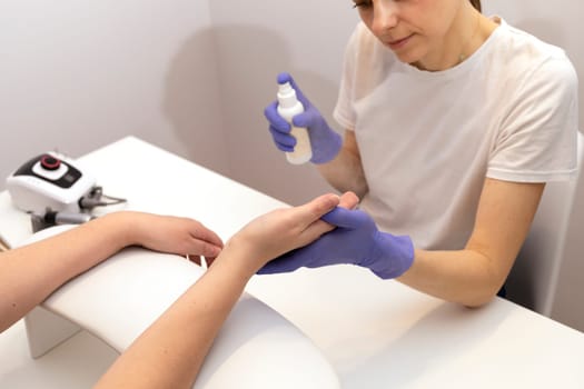 Nails master spraying, sanitizing client's hands by antiseptic from white bottle at work station. Beautician wears gloves, holding woman's hand over table. Hardware nail machine is beside.Horizontal